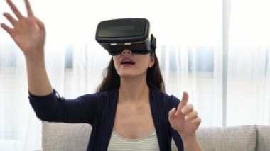 Dolly atış genç kadının koltukta oturur ve eğleniyor Vr kulaklık gözlükle eller yukarı evde oturma odasında oyun oynama.