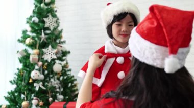 güzel Asya anne yay kravat ve Noel Baba gibi birlikte evde oturma odasında giyinmek için mutlu sevimli küçük Asyalı çocuk kızı yardımcı olur. Kış tatil tatil kavramı.