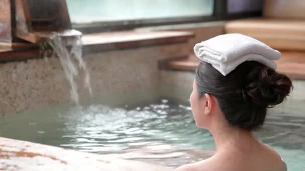 年轻女孩的后景色享受温泉 并在她的头上放一条毛巾 而水在背景上流动 — 图库视频影像