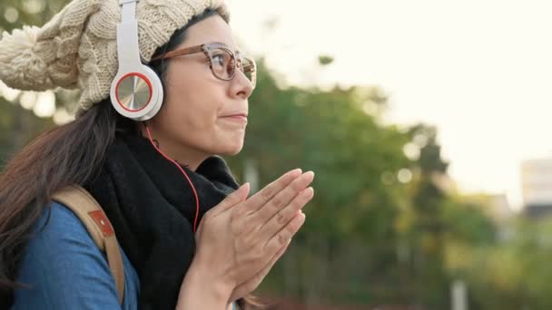 美しいアジアの少女をこする彼女の手と摩擦熱に暖かい都市公園で音楽を楽しむ — ストック動画