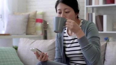 Asyalı kadın Smartphone cep telefonu ile mesaj okuma oturma odasında kahve içiyor iken.