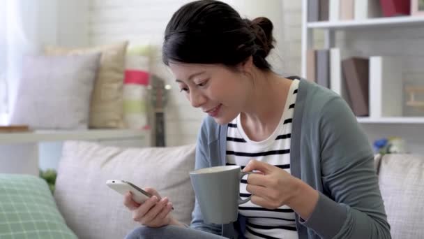 彼女は携帯電話のテキスト メッセージ生活や飲み物のコーヒーのソファに座っているアジア女性の早送り — ストック動画
