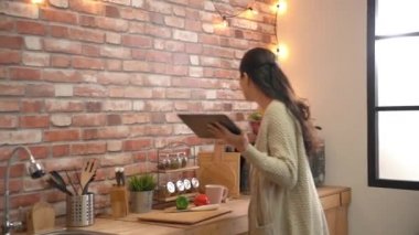 Ağır çekim bir kadın tarafından tablet online dijital tarifi arıyorum. O şarkı ve bir gülümseme ile mutfakta dans çok sevindi. Pişirmek hazırlar.