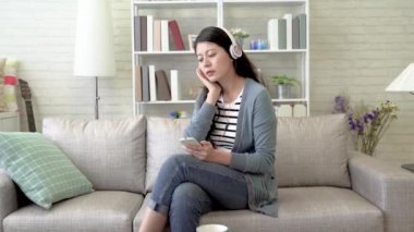 Asya genç online cep telefonu ve kulaklık ile müzik dinlemek. O oturma odasında kanepede oturuyor ve melodi takip şarkı sözleri şarkı söylüyor.