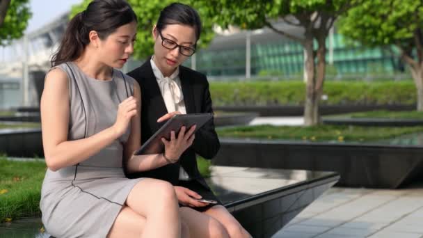 两位亚洲妇女讨论与数字平板设备坐在办公室公园外面与伟大的阳光天气天 — 图库视频影像