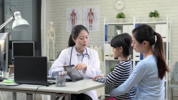 亚洲妇女医生和小女孩和妈妈使用数字平板电脑垫在诊所一起讨论 — 图库视频影像