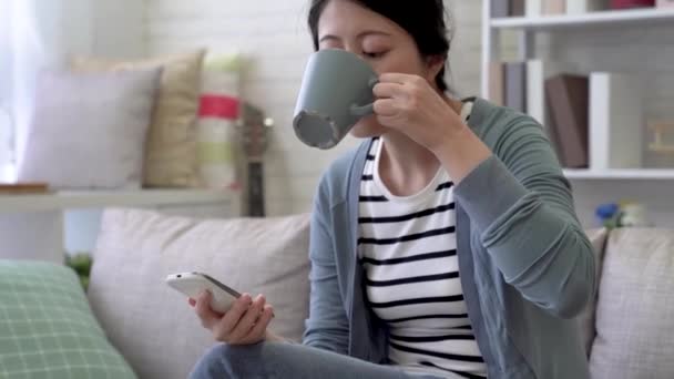 アジアの女性は 彼女は彼女が携帯電話を使用してリビング ルームでそれを飲んだ後 コーヒーに肯定的な認識を与えた — ストック動画