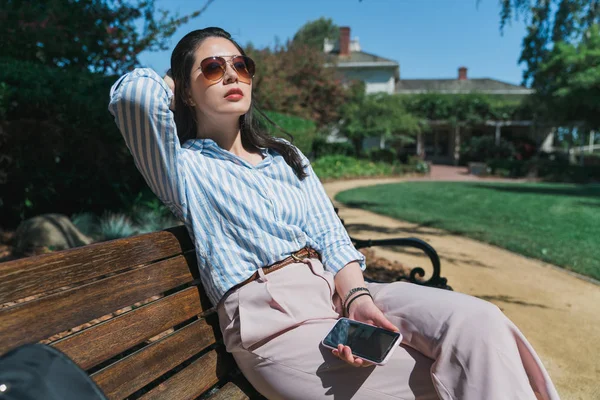 Mulher glamourosa usando óculos de sol sentar no banco Imagem De Stock