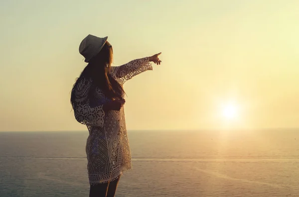 Mujer joven con sombrero y lindo vestido de verano de pie sobre la piedra, mirando el atardecer sobre el mar — Foto de Stock