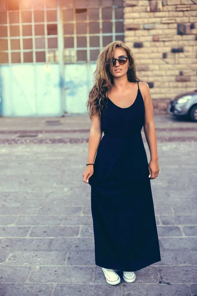 Mujer bonita joven en vestido negro, gafas de sol y zapatillas blancas al atardecer en Italia . — Foto de Stock