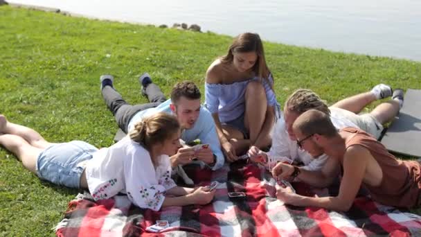 Jonge mensen zittend op het groene gras en speelkaarten in de buurt van het meer. — Stockvideo