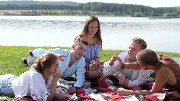 Jóvenes sentados en la hierba verde y jugando a las cartas cerca del lago . — Vídeos de Stock