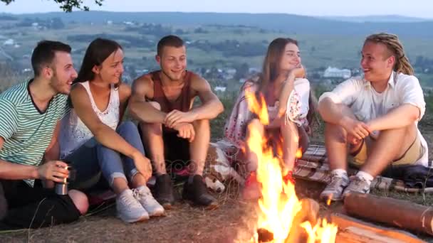 Groep vrienden zitten naast een kampvuur met warme dranken en praten — Stockvideo