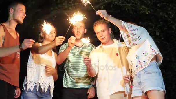 Jóvenes alegres sentados el fin de semana por la noche con chispas de fuegos artificiales . — Vídeo de stock