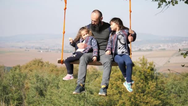 Papai balançando com filhas em um balanço debaixo de uma árvore . — Vídeo de Stock