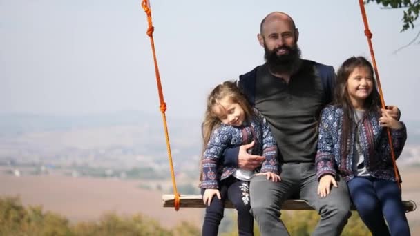 Retrato de papá balanceándose con sus hijas en un columpio bajo un árbol . — Vídeo de stock