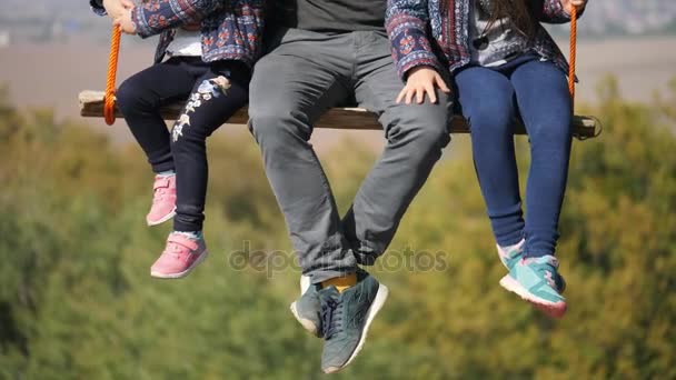 Ноги отца, качающегося с дочерьми на качелях под деревом. Крупный план — стоковое видео