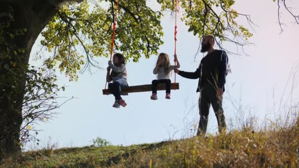 Papá sacude a sus hijas en un columpio bajo un árbol . — Vídeo de stock