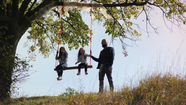 Papa schüttelt Töchter auf Schaukel unter Baum. — Stockvideo