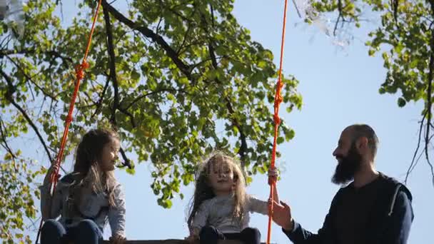 Papá sacude a sus hijas en un columpio bajo un árbol . — Vídeo de stock