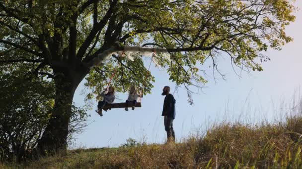 Papa schudt haar dochters op een schommel onder een boom. — Stockvideo