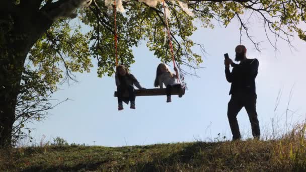 Padre fare foto delle sue due figlie che oscillano su un'altalena sotto un albero . — Video Stock