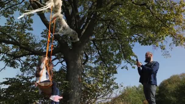 Père faire la photo de ses deux filles se balançant sur une balançoire sous un arbre . — Video