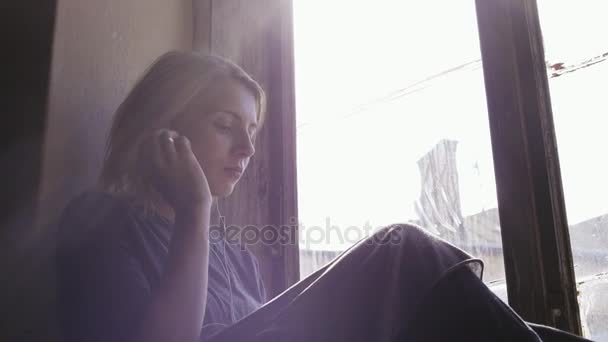 女の子は非常に悲しそうに見える一方、窓のそばのヘッドホンで音楽を聴く。クローズ アップ — ストック動画