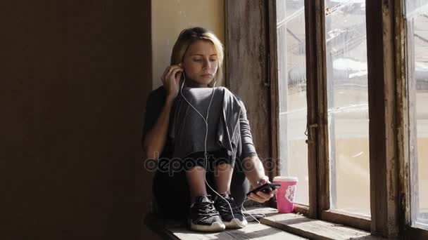 女の子は窓のそばにヘッドフォンで音楽を聞いている間に非常に悲しいように見えます — ストック動画