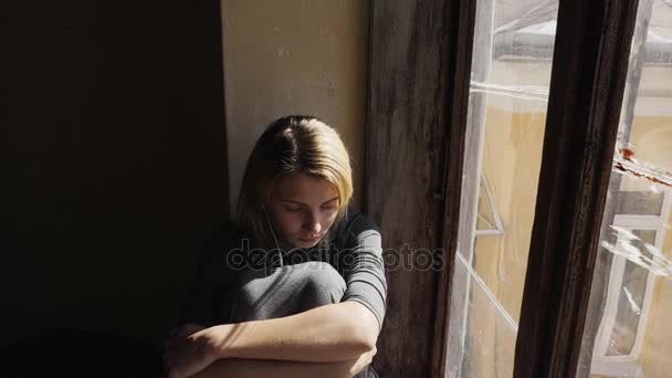 女の子は窓のそばにヘッドフォンで音楽を聞いている間に非常に悲しいように見えます — ストック動画