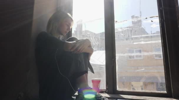 Chica se ve muy triste mientras escucha música en los auriculares por la ventana — Vídeos de Stock