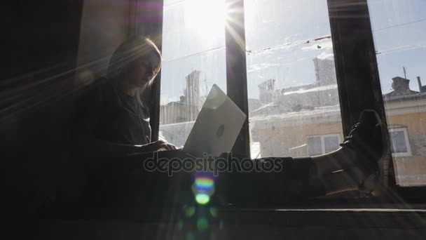 悲しい女の子はヘッドフォンで音楽を聞いて窓のそばに座って、ラップトップを使用して — ストック動画