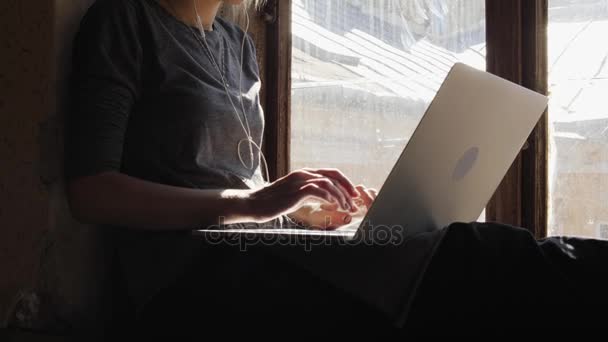 Chica triste sentado junto a la ventana escuchando música en los auriculares y utilizando un ordenador portátil — Vídeos de Stock