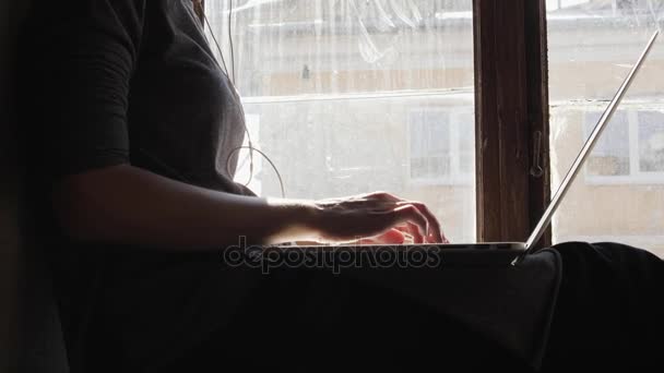 Chica triste sentado junto a la ventana escuchando música en los auriculares y utilizando un ordenador portátil — Vídeos de Stock