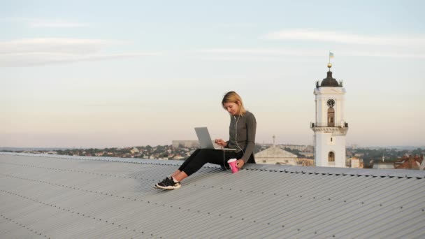 Junge Frau sitzt mit Laptop und Musik über Kopfhörer auf dem Dach. — Stockvideo