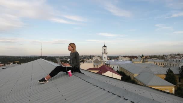 Jeune femme est assis avec ordinateur portable et écouter de la musique sur les écouteurs sur le toit. — Video