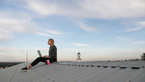 Ung kvinna sitter med laptop och lyssnar musik på hörlurar på taket. — Stockvideo