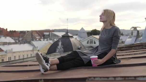Jonge gelukkige vrouw zit met koffie op het dak. Wind waait haar haren. — Stockvideo