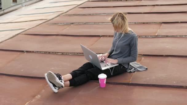 Молода жінка сидить з ноутбуком і слухає музику на навушниках на даху . — стокове відео