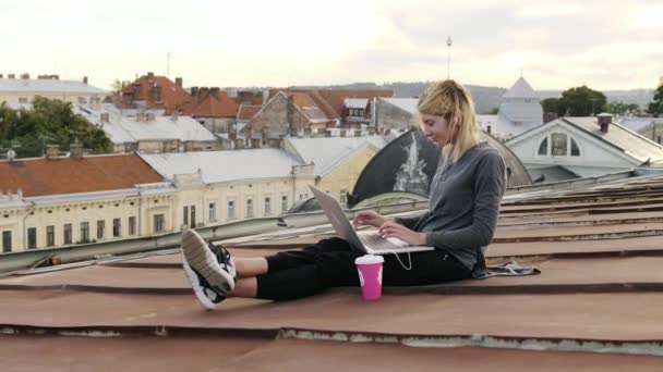 Mladá žena sedí s laptopem a poslouchá hudbu na sluchátkách na střeše. — Stock video