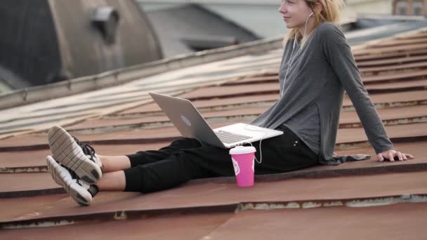 Jonge vrouw zit met laptop en muziek te luisteren op de hoofdtelefoon op het dak. — Stockvideo