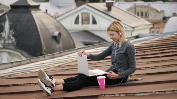 Молодая женщина сидит с ноутбуком и слушает музыку на наушниках на крыше. — стоковое видео