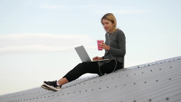 Young woman sits with laptop and listening music on headphones on the rooftop. — Stock Video