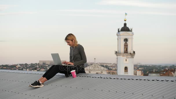 Молода жінка сидить з ноутбуком і слухає музику на навушниках на даху . — стокове відео