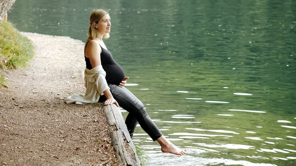 Vacker, söt gravid kvinna sitter nära en fjällsjö i en skog på stranden — Stockfoto