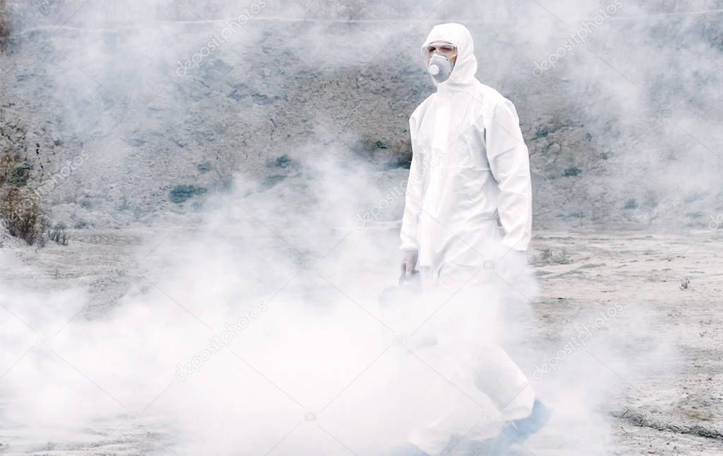 Lab technician in a mask and chemical protective suit, walks on dry ground with a tool box through toxic smoke