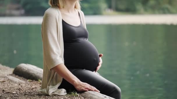 Zwangere vrouw die zachtjes haar buik aanraakt als ze bij het bergmeer zit. — Stockvideo