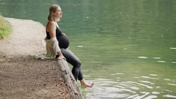 Mujer embarazada sentada en el borde de una tabla de madera en el lago y balanceando los pies en el agua . — Vídeos de Stock