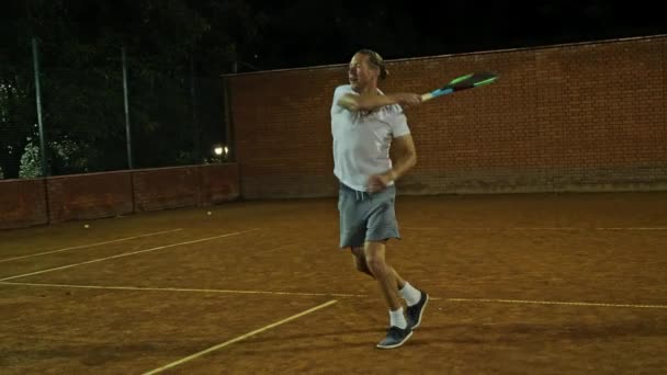 Spelar tennis på natten. Ung attraktiv man spelar tennis på orange lera tennisbana — Stockvideo