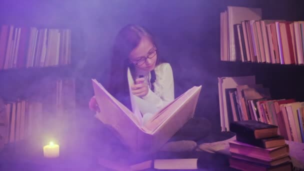 Ein Mädchen schlägt in der Bibliothek ein Märchenbuch auf, farbiger Rauch wirbelt herum, Kerzen brennen in der Nähe — Stockvideo